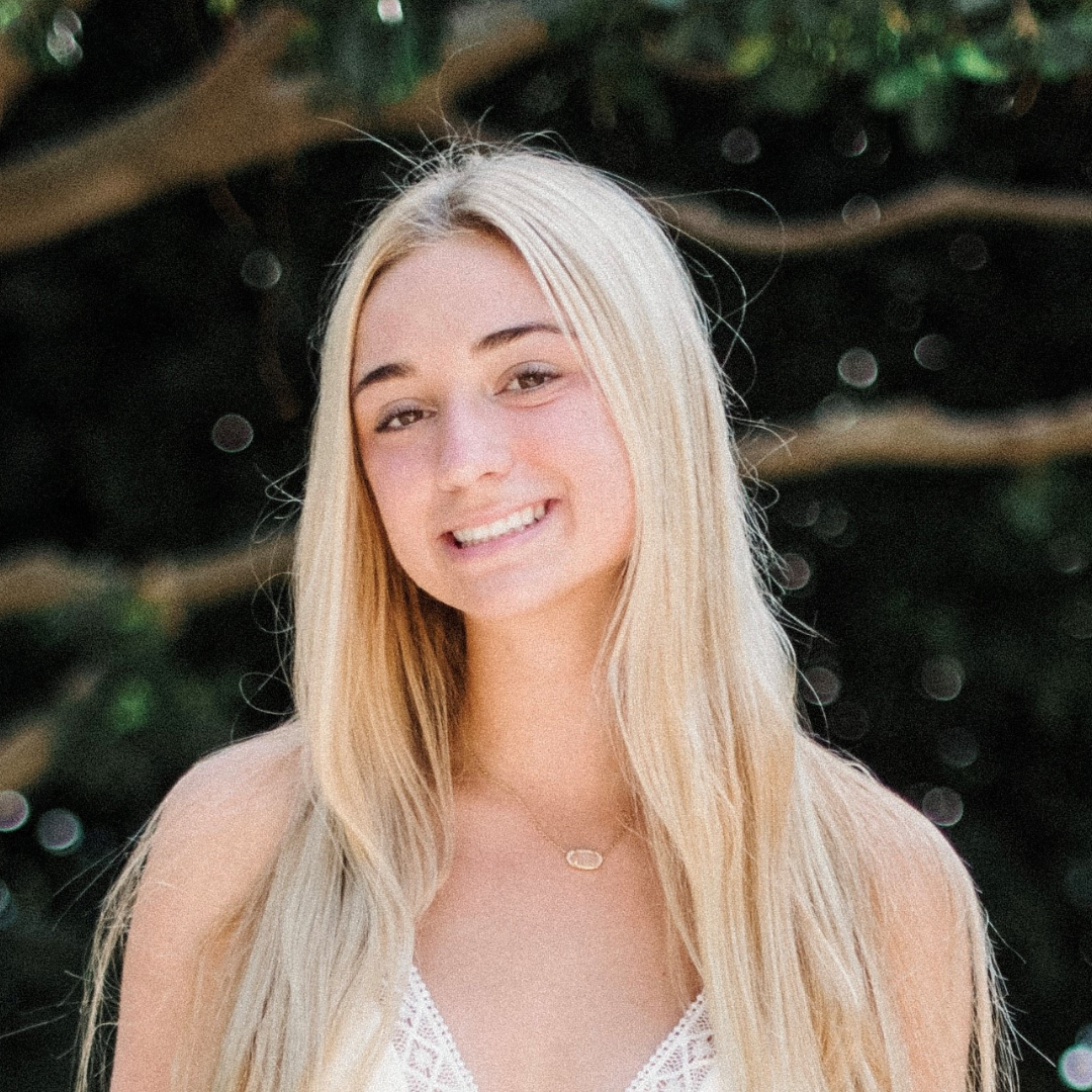 A woman with long blonde hair and wearing a white tanktop is smiling at the camera.
