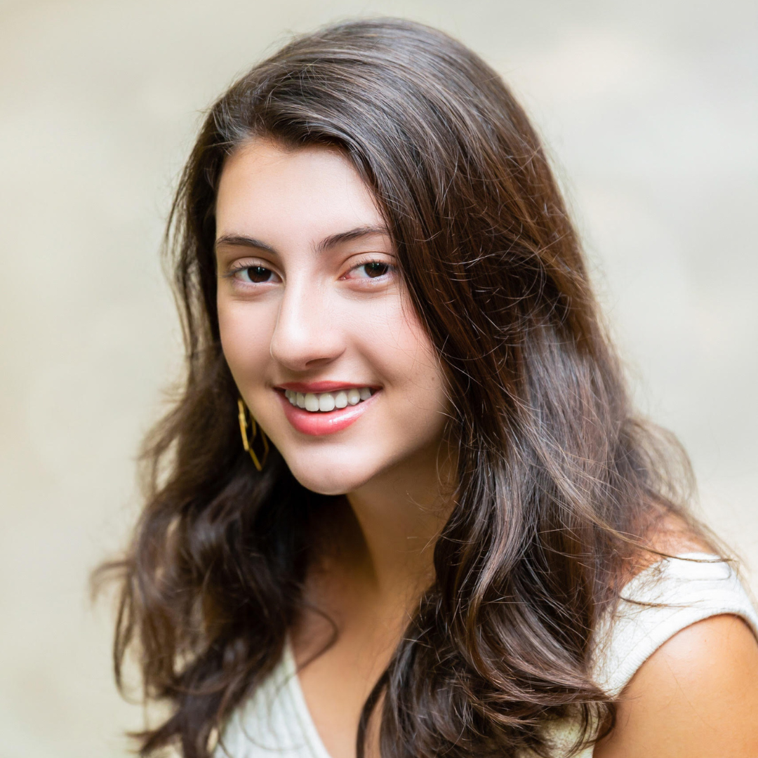 A woman with long, brown curled hair and brown eyes is smiling at the camera and wearing a white blouse and gold, hoop earrings