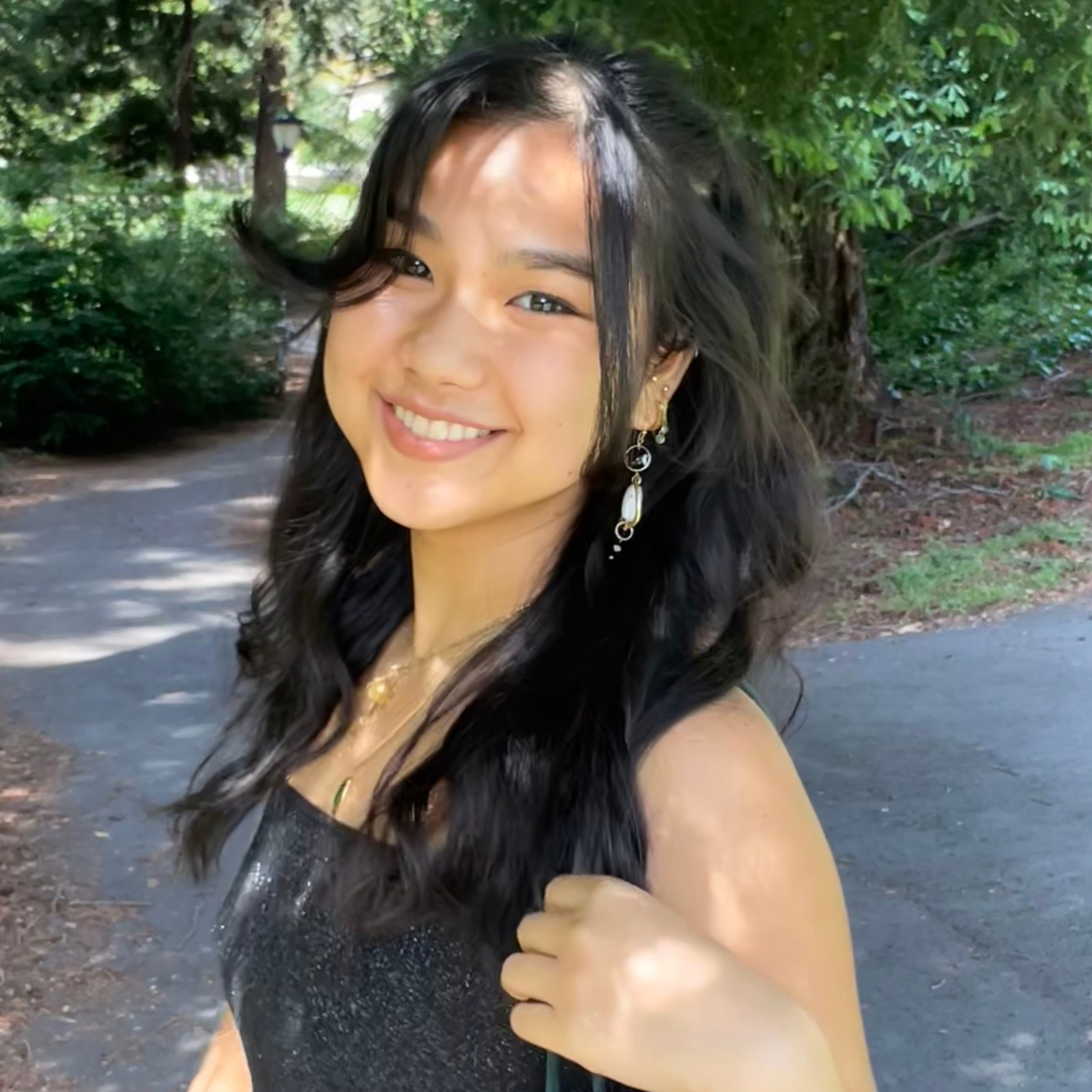 A woman with long, wavy black hair and brown eyes is turned and smiling at the camera while wearing a black strapless top and pearl earrings