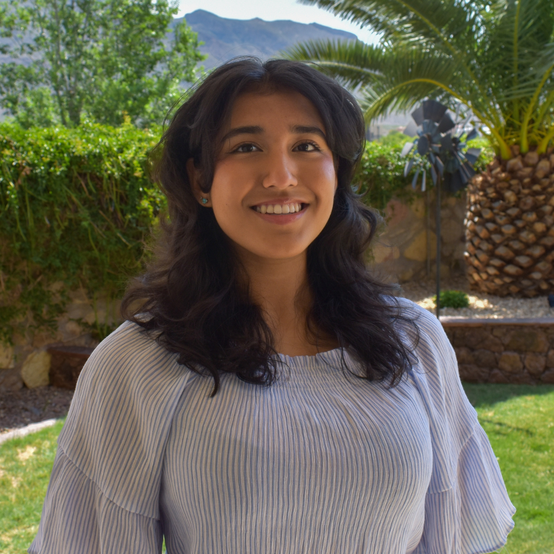 A woman with medium-length, slightly curly black hair is looking at the camera and smiling. She is wearing a top with elbow-length sleeves that's ruffled.