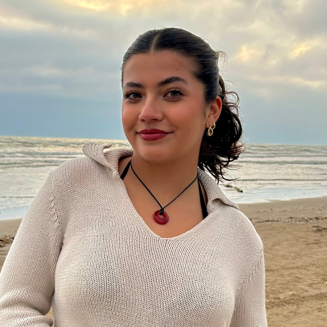 A young woman is seen on a beach. She has dark brown hair tied up and is wearing a cream sweater.