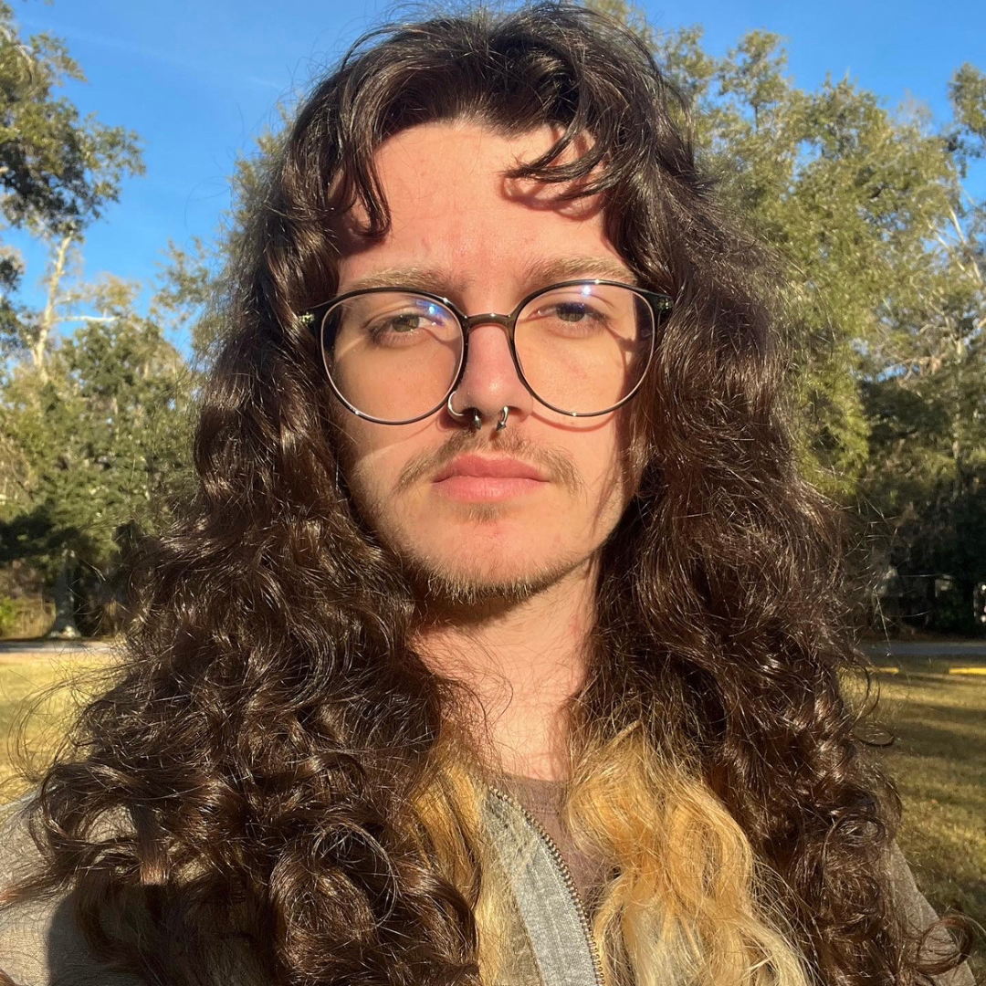 A male-presenting student with glasses and long, curly, brown hair is seen looking into the camera.
