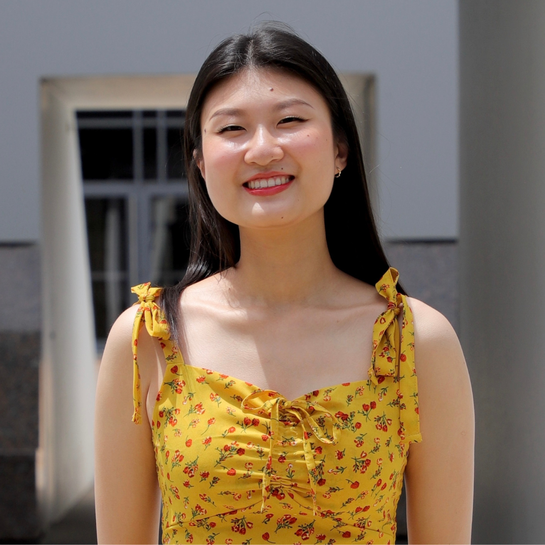 A woman with long, straight, black hair is standing and smiling at the camera while wearing a yellow and red floral dress