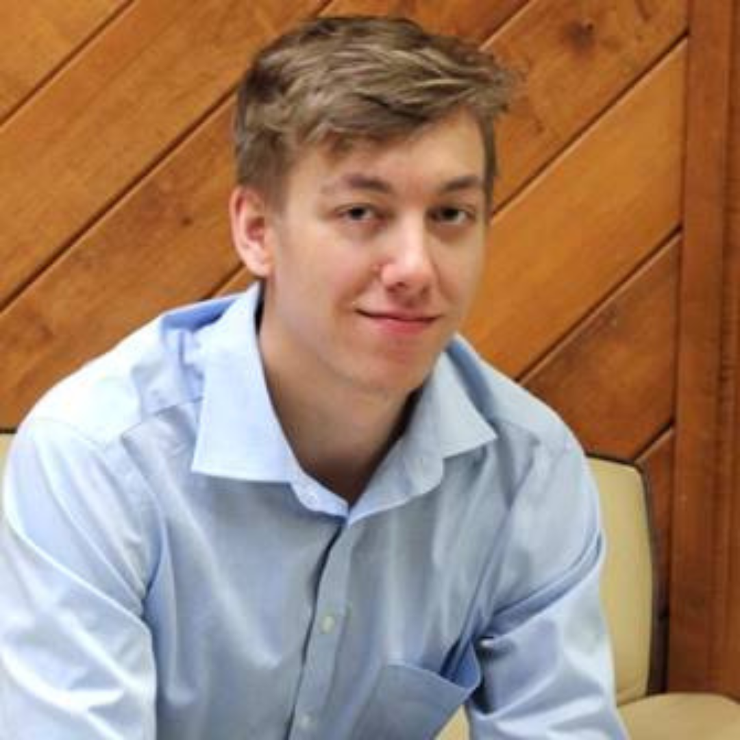 A man with short, blond hair is seen smiling at the camera and wearing a blue button-down shirt