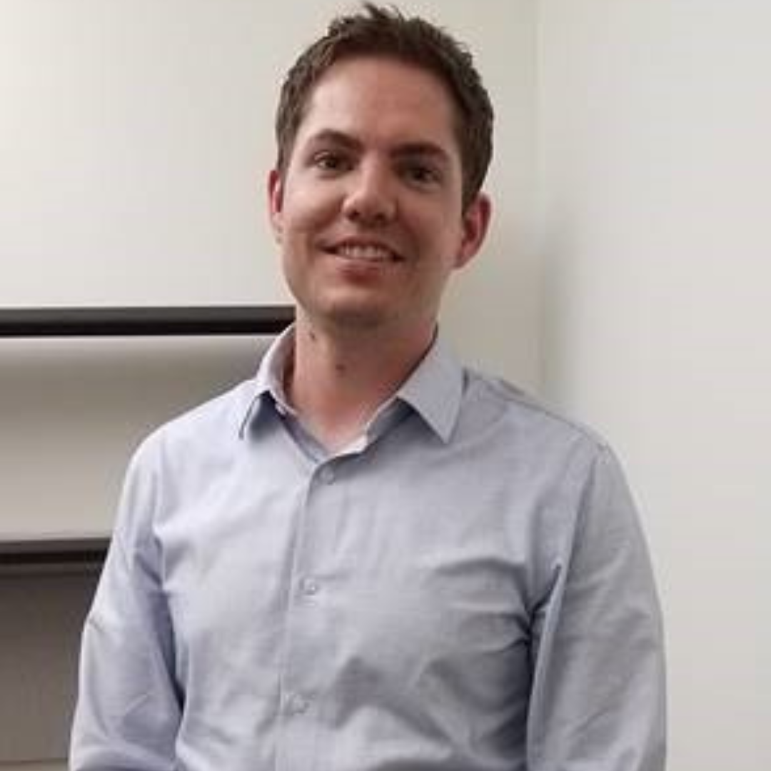 A man with short, brown hair is standing and smiling at the camera wearing a light blue button-down shirt