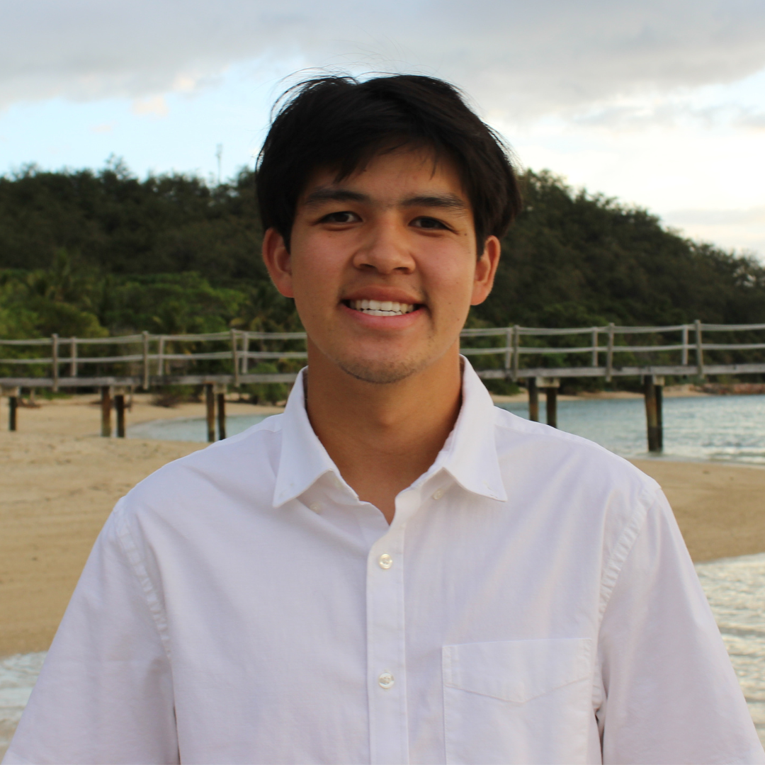 A man with ear-length, straight black hair is wearing a white button-up shirt and smiling on the camera while standing at the beach