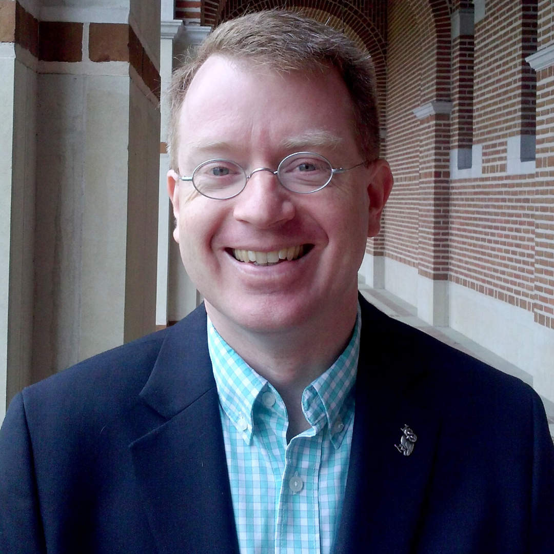 A man with short, red hair and glasses is seen smiling at the camera and wearing a navy suit jacket