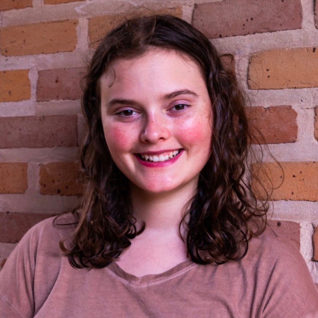 A woman with shoulder-length, curly brown hair is smiling at the camera. She is wearing a brown t-shirt and standing in front of a brick wall.