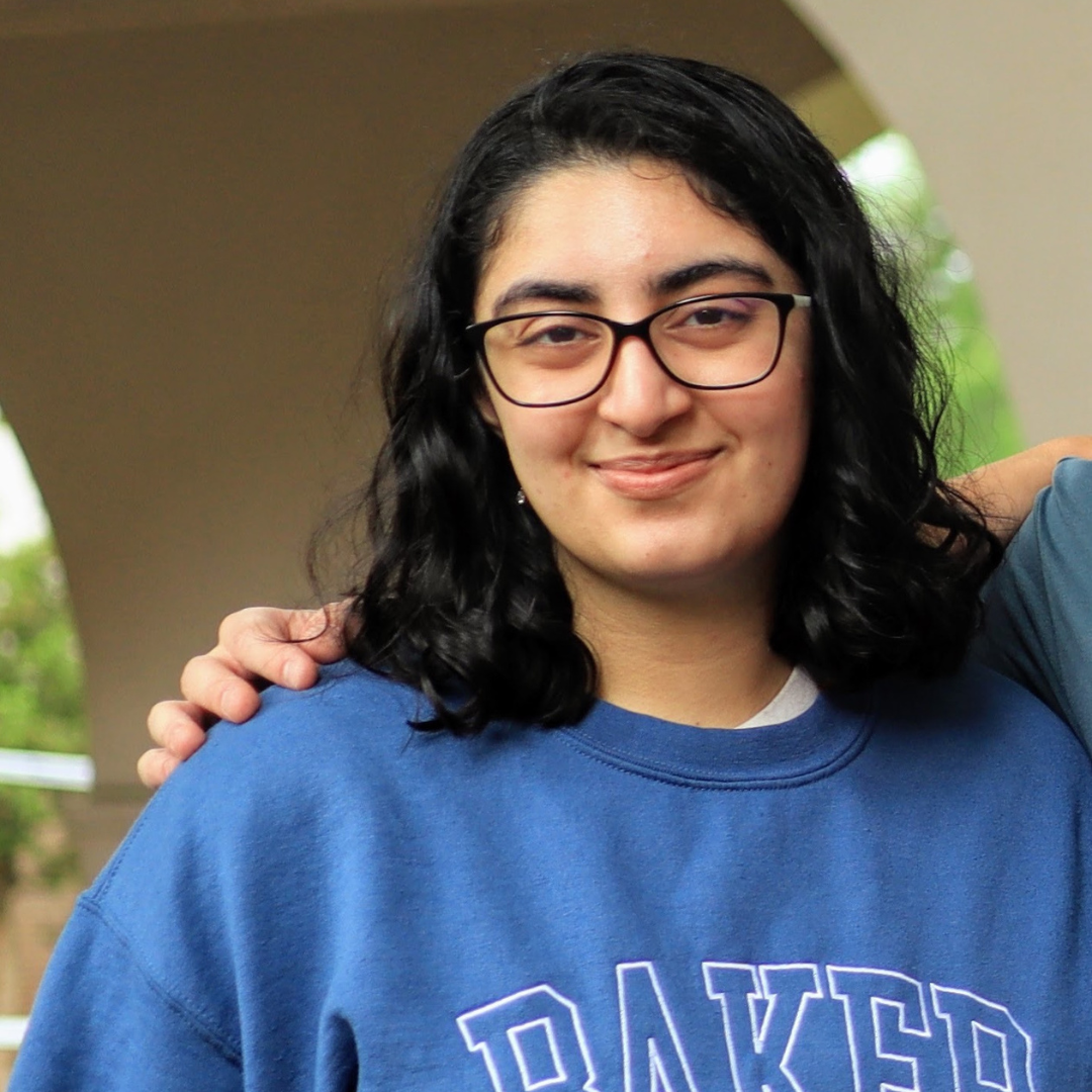 A woman with shoulder-length, curly black hair and brown eyes is wearing glasses and a blue sweater with the word "Baker" across it