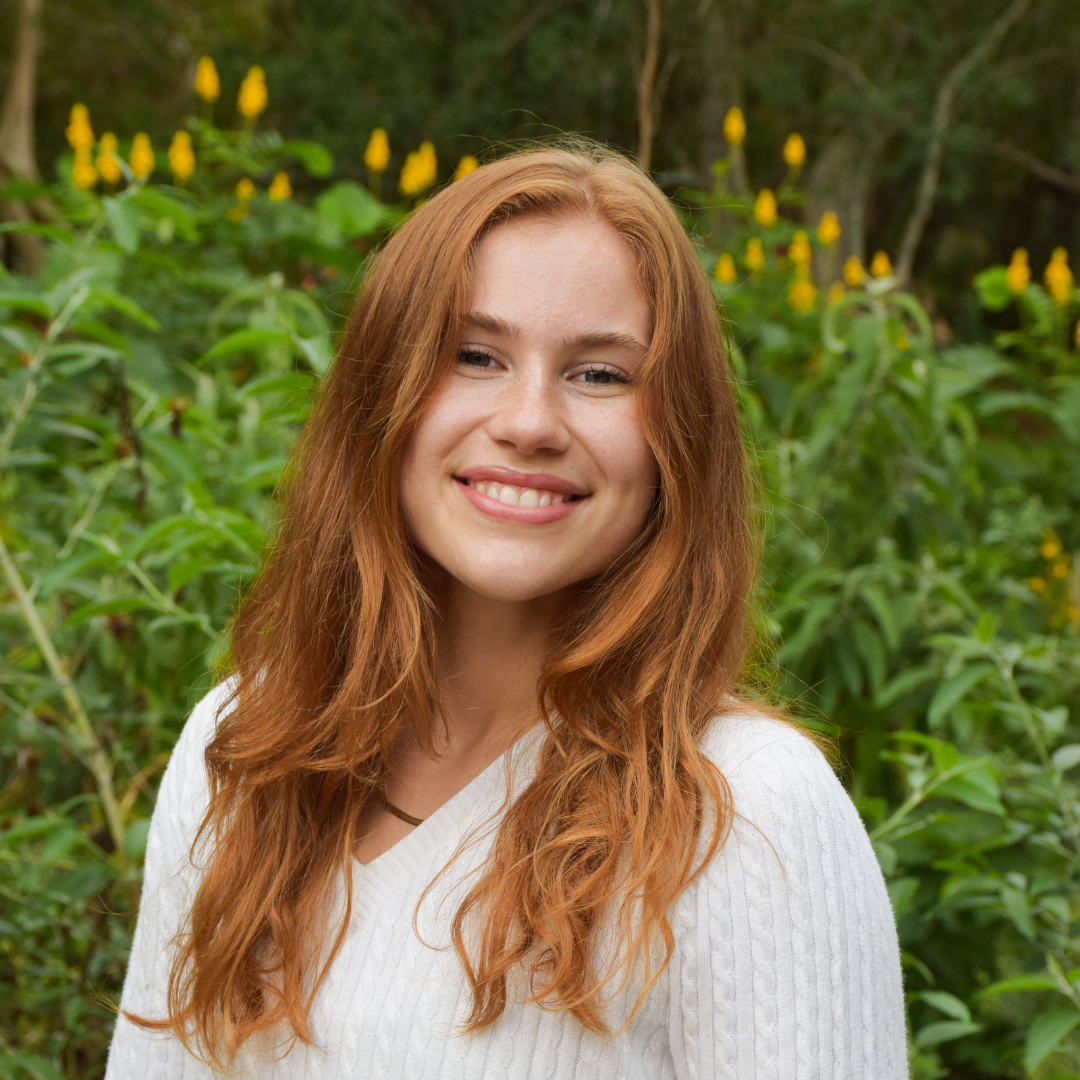 A woman with long, curly red hair is smiling at the camera and wearing a white cable-knit sweater.