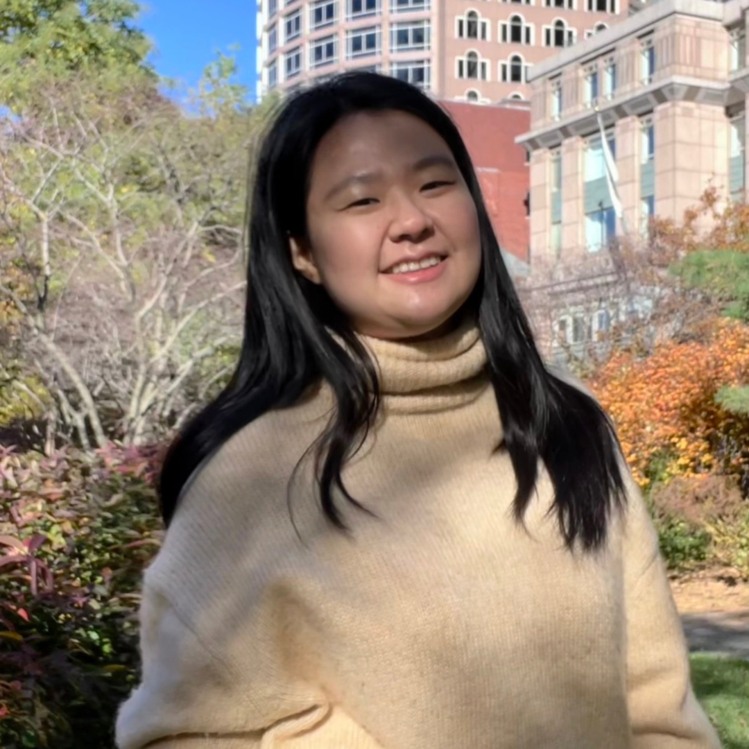 An Asian, female student is seen outside. She is wearing a cream-colored sweater and has straight, shoulder-length black hair.