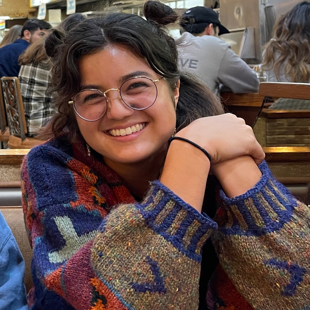 A woman with short, curly brown hair and brown eyes is wearing glasses, a sweater and smiling at the camera
