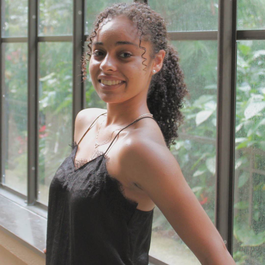 A woman with super curly brown hair is wearing a black tank top and smiling at the camera while leaning against a glass wall