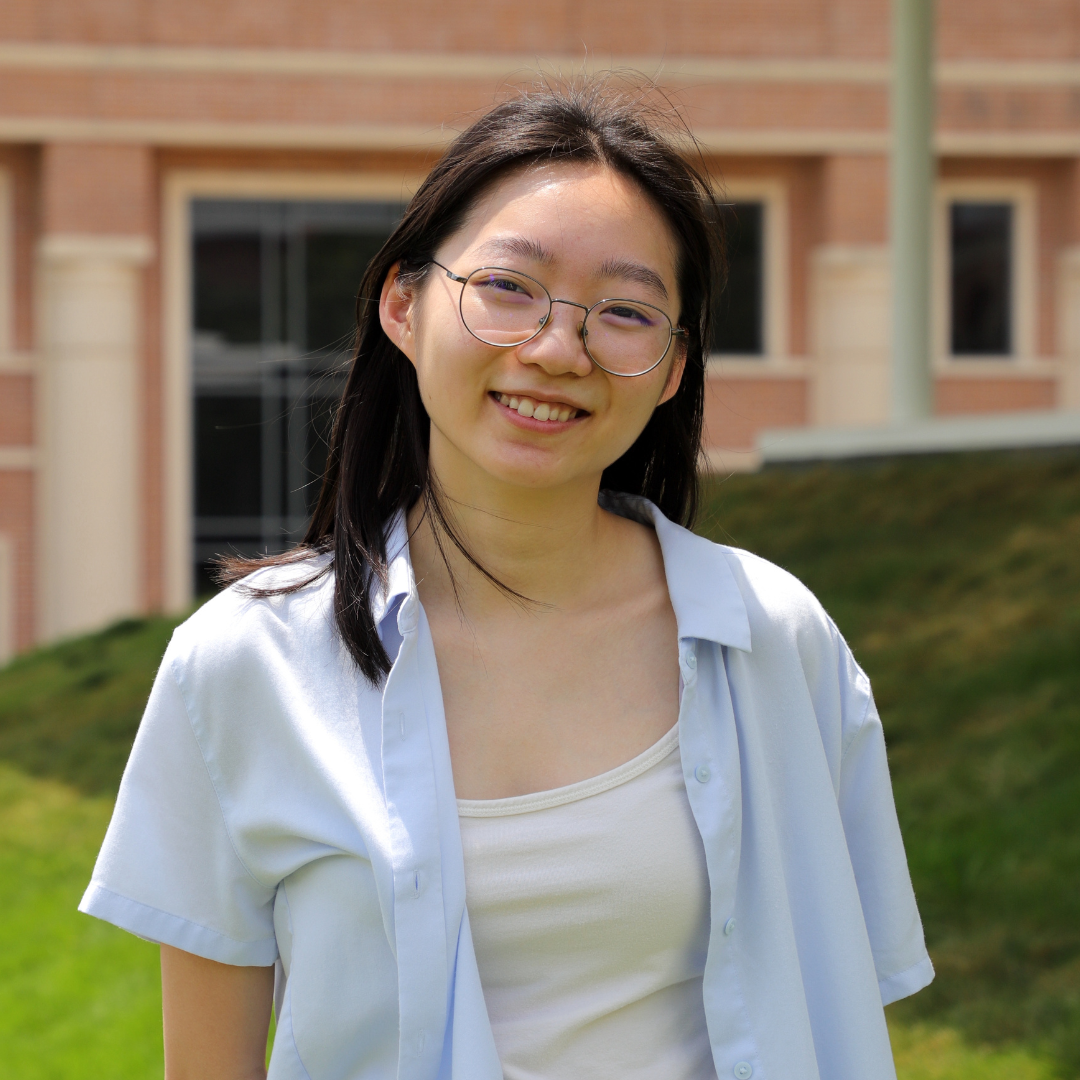 A woman with shoulder length, straight black hair is wearing glasses and a light blue shirt over a white tank top
