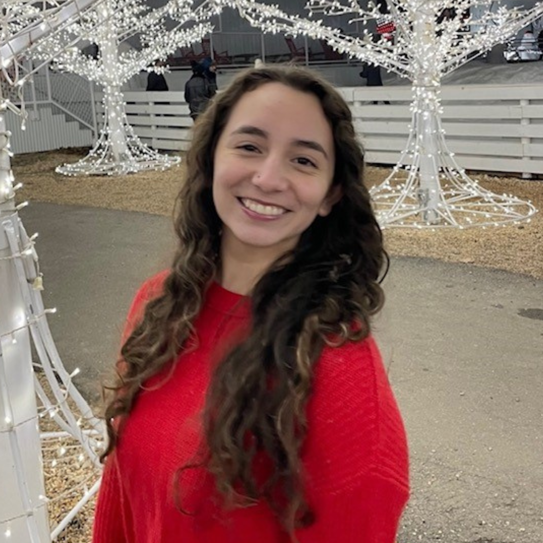 A woman with long, brown, curly hair is smiling at the camera. She is wearing a red sweater and standing in front of trees made of white lights.