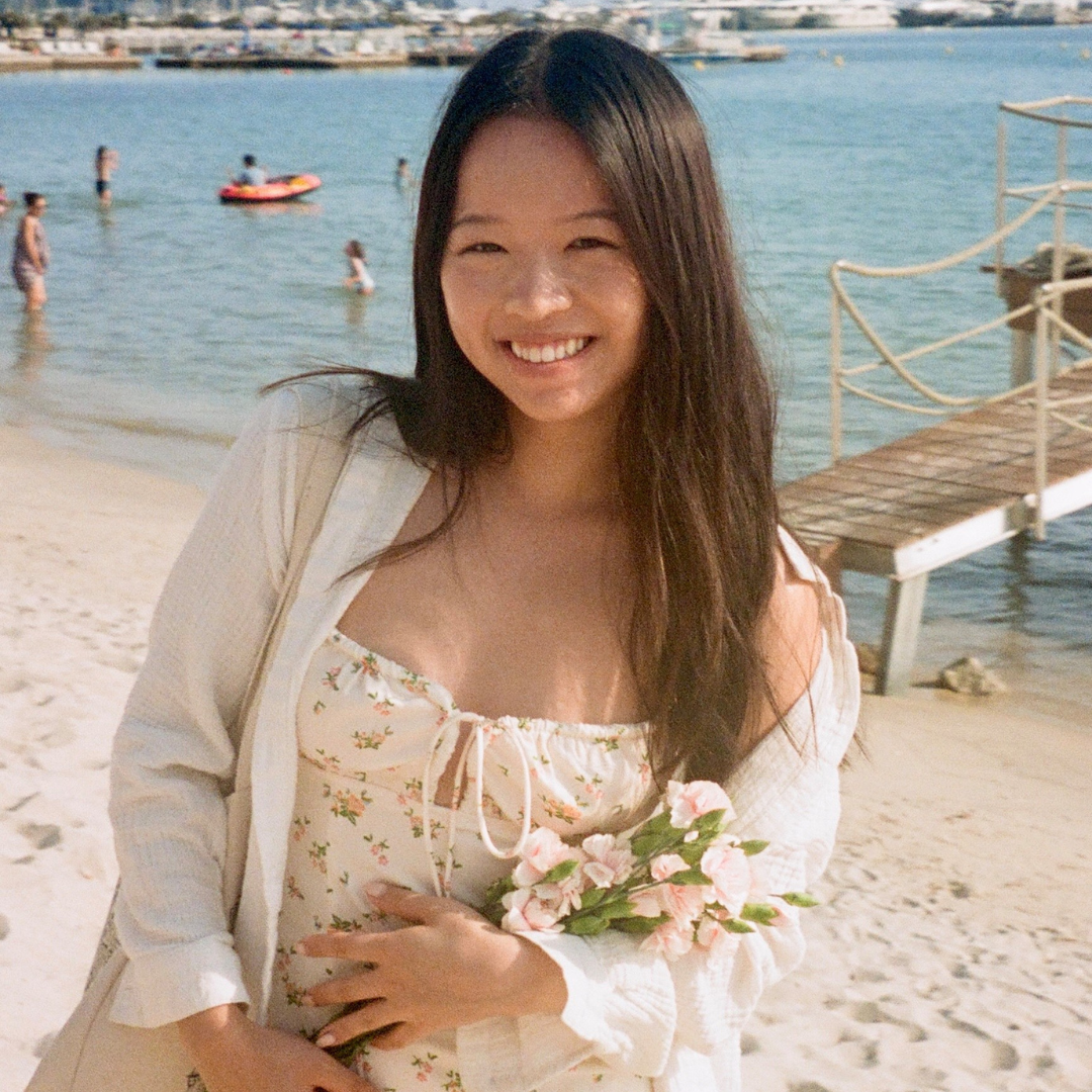 A young woman is seen on a beach. She has long, dark brown hair and is wearing a cream, floral dress.