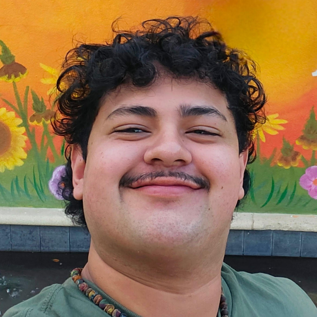 A male student with curly, black hair and a mustache is smiling at the camera. He is wearing a green t-shirt.