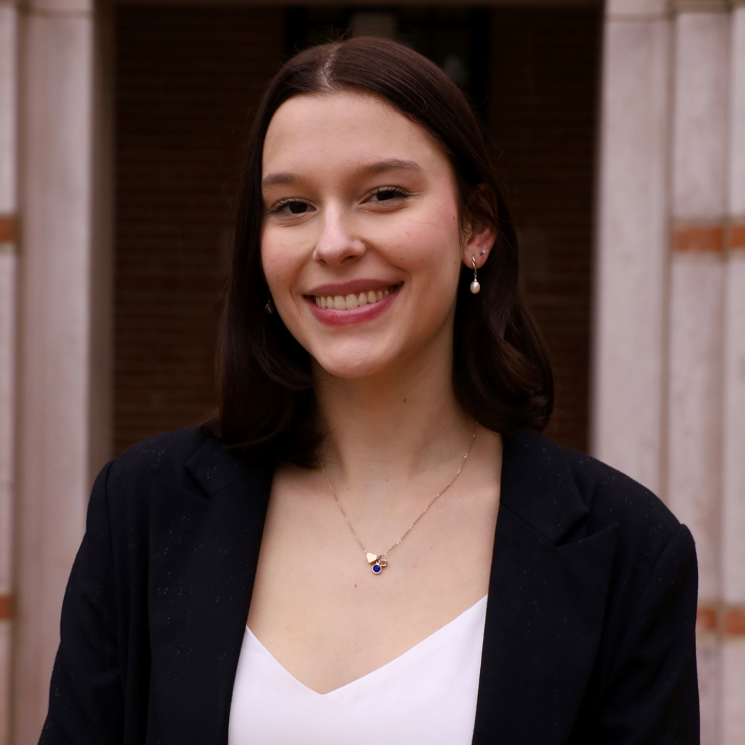 A female student with shoulder length, dark brown hair and brown eyes is seen smiling at the camera. She's wearing a white blouse with a black blazer.