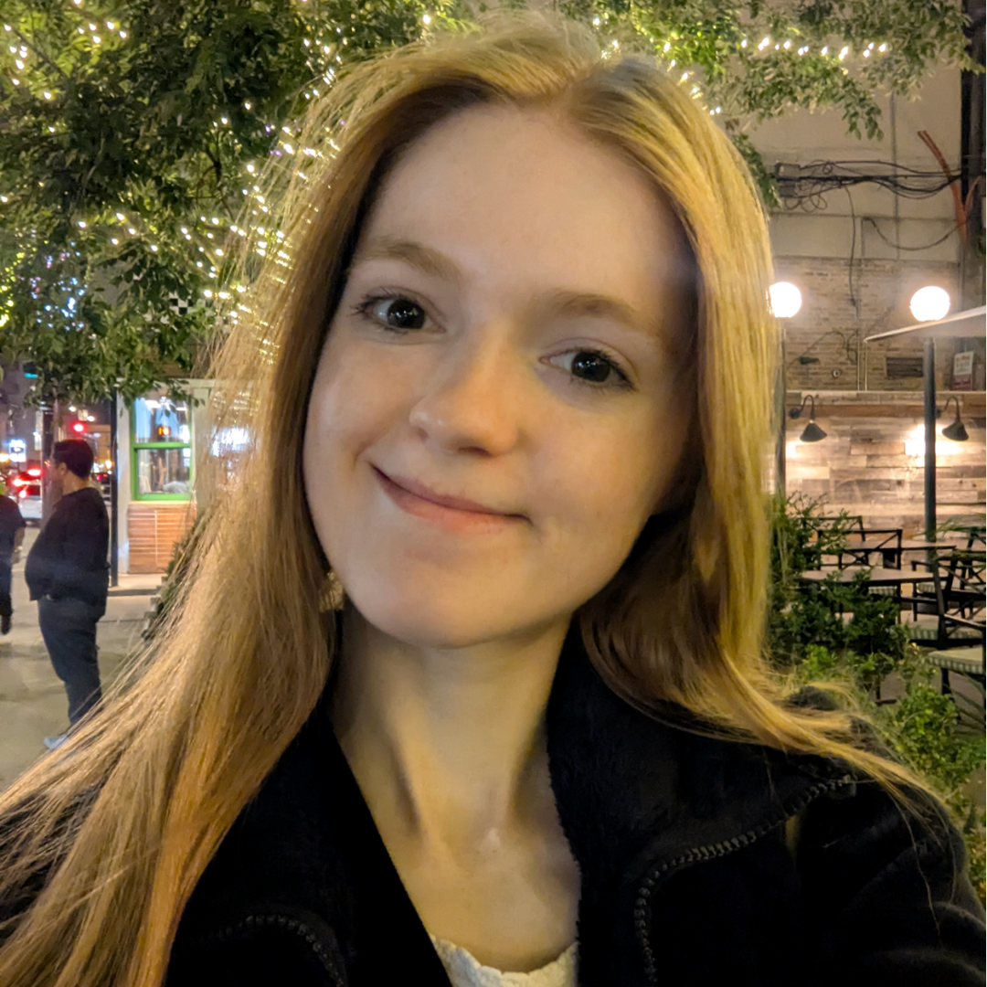 A female student is staring into the camera with a small smile. She has long, reddish-blonde hair, brown eyes, and is wearing a zipped up black jacket.