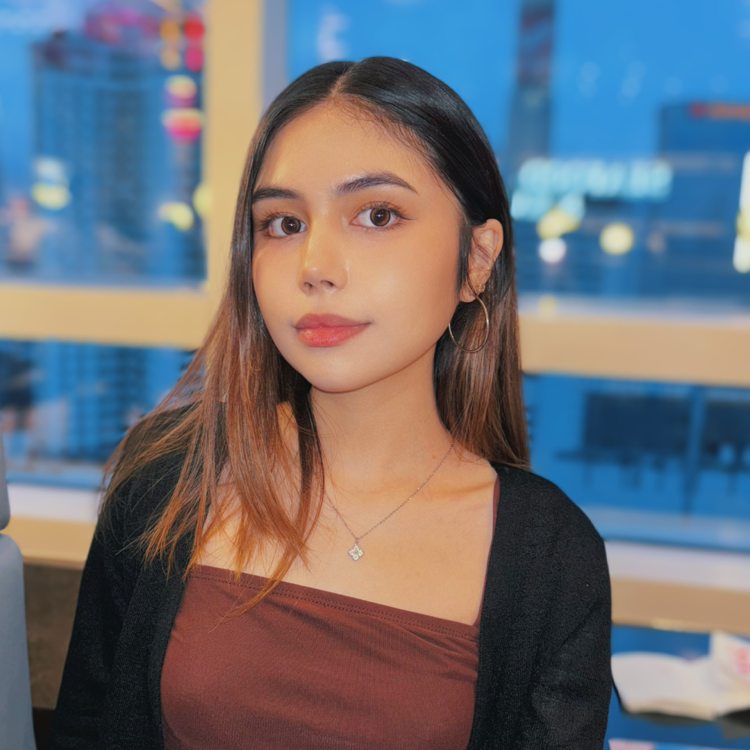A female student with long, ombre black to brown hair is looking at the camera. She's wearing a brown tank top and a black cardigan.