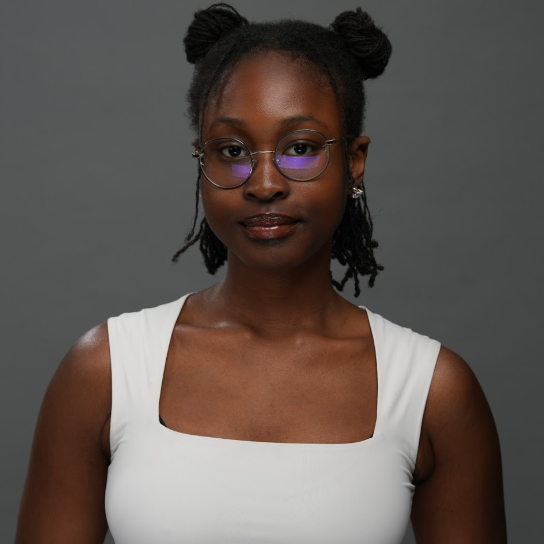 A female student is staring into the camera with a slight smile. Her shoulder-length black hair is partially up in space buns. She has glasses and is wearing a white, square-neck top.