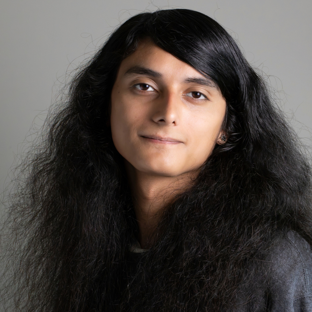 A male student with long, wavy black hair is smiling slightly at the camera. He is wearing a gray sweatshirt.