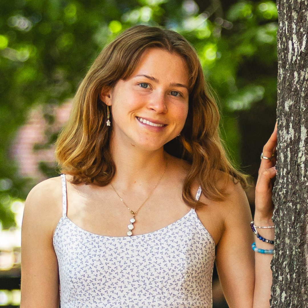 A female student with shoulder length, wavy dark blonde hair is smiling at the camera while leaning against a tree and waring a white dress with a small floral pattern