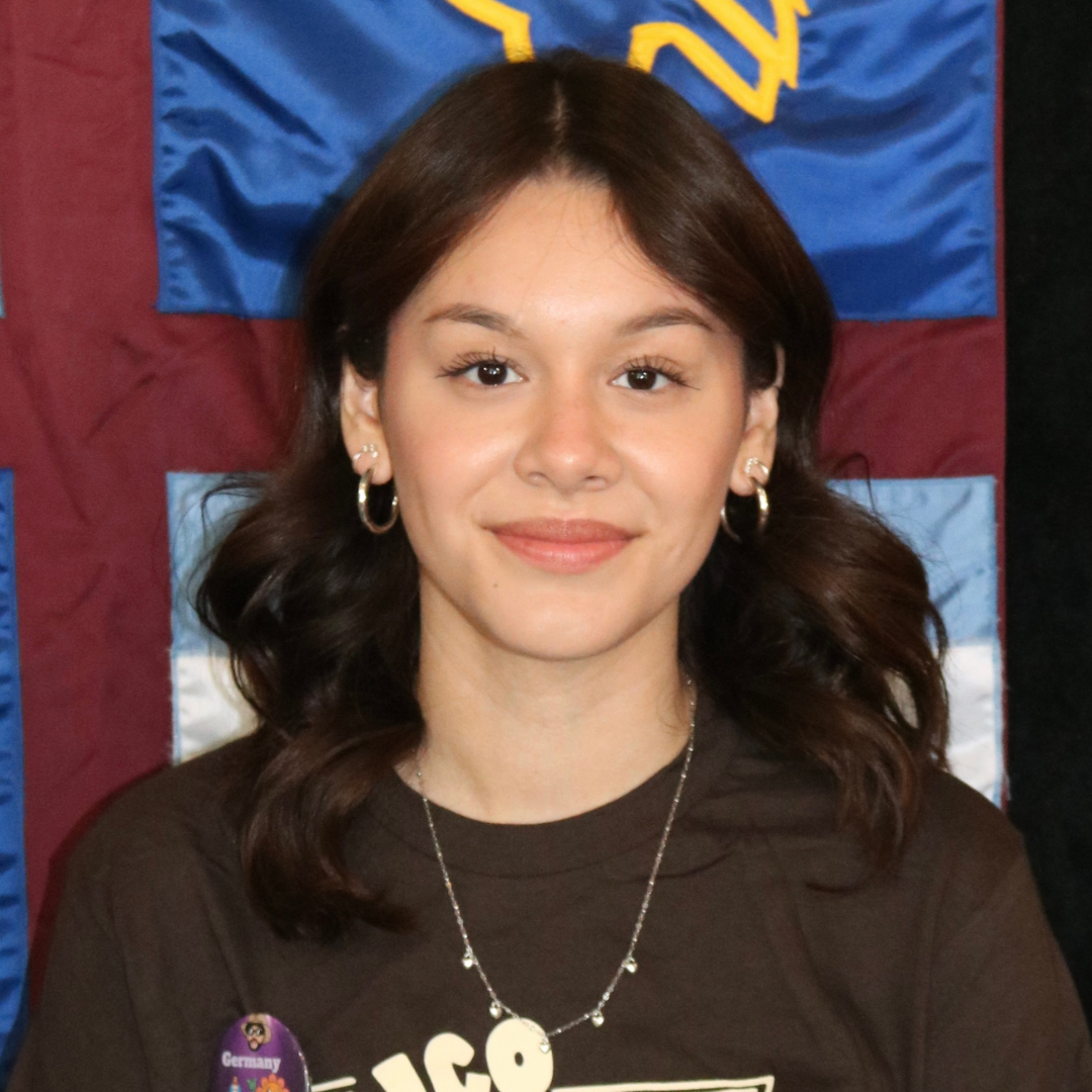 Image of a female student with short, wavy dark brown hair is smiling at the camera and standing in front of a Martel College banner