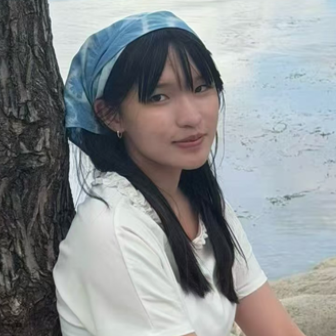A female student is leaning against a tree in front of water. She's wearing her long, black hair down with a blue bandana tied around her head.