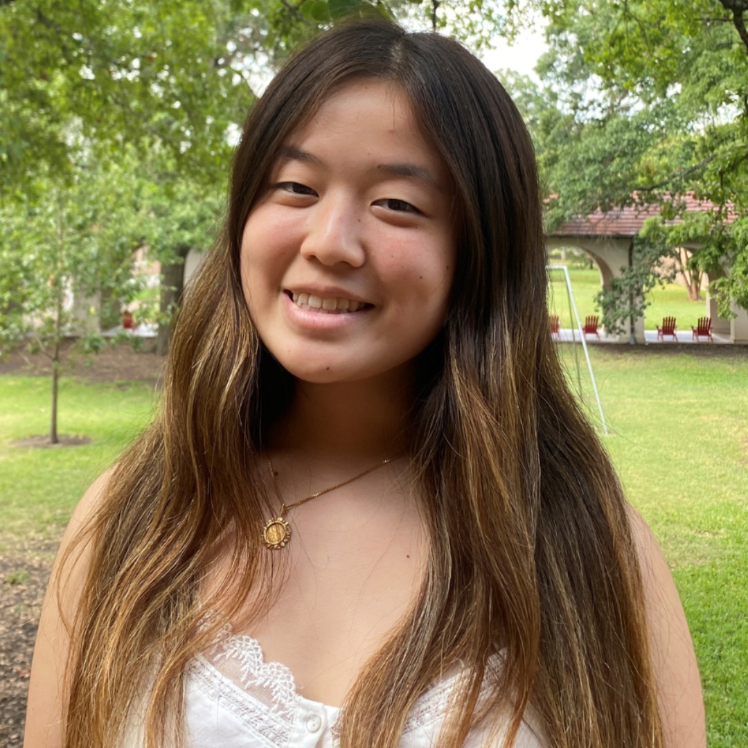 A female student with long, ombre black to brown hair is looking at the camera. She's wearing a brown tank top and a black cardigan.