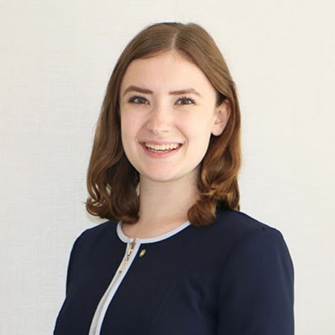 A female student with short, curled, light brown hair is smiling at the camera and wearing a white and navy suit jacket