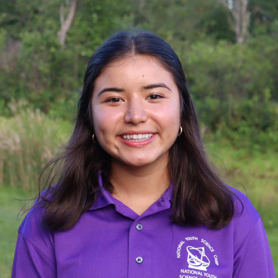 A female student with long, dark brown hair is smiling at the camera and wearing a purple polo