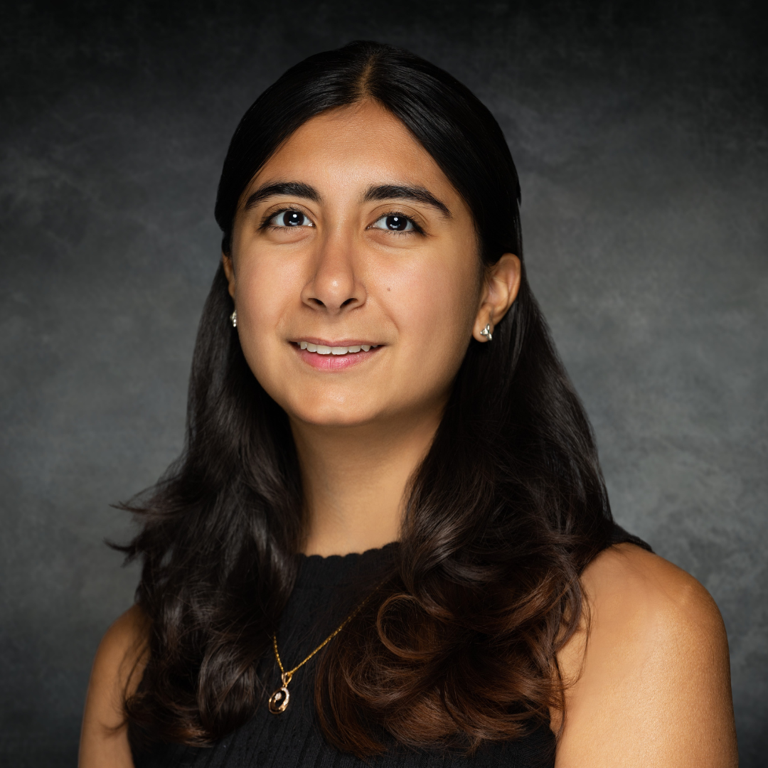 A female student with long, dark brown curly hair is shown looking into the camera and smiling. She is wearing a black, sleeveless blouse and gold necklace.