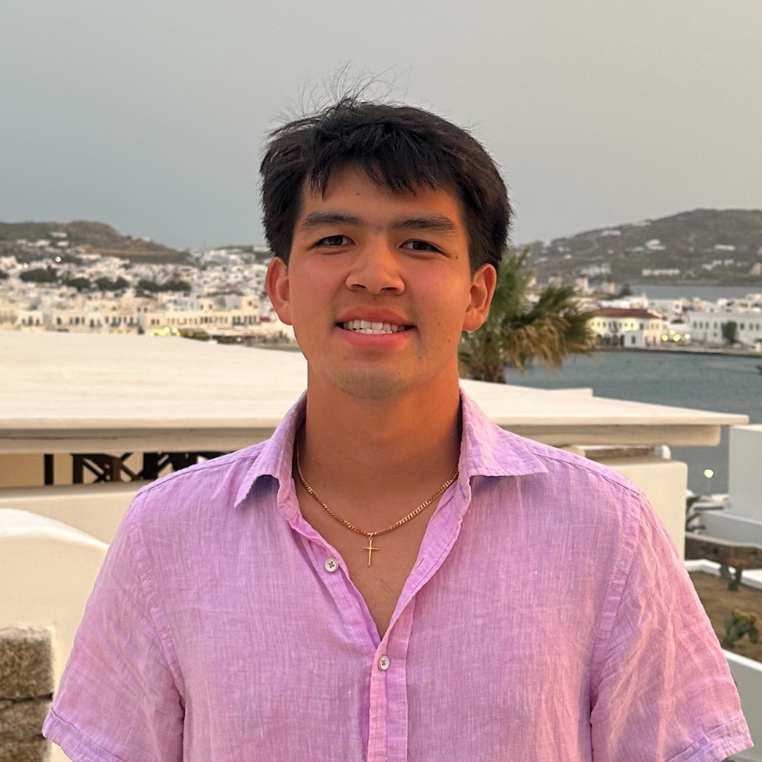 A male student with ear length black hair is smiling at the camera. He is wearing a pink, button-up shirt