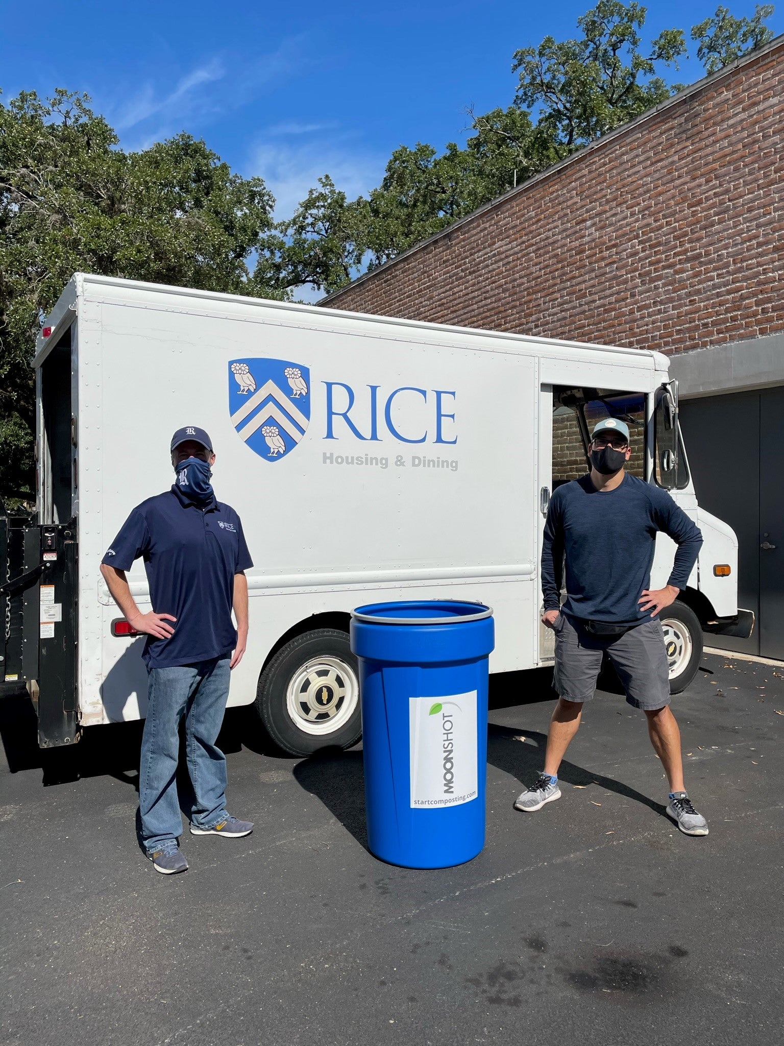 Two people stand outdoors next to a large blue compost bin labeled “Moonshot Compost” in front of a white truck with the Rice University Housing & Dining logo. The truck is parked beside a brick building, and trees are visible in the background under a clear blue sky.