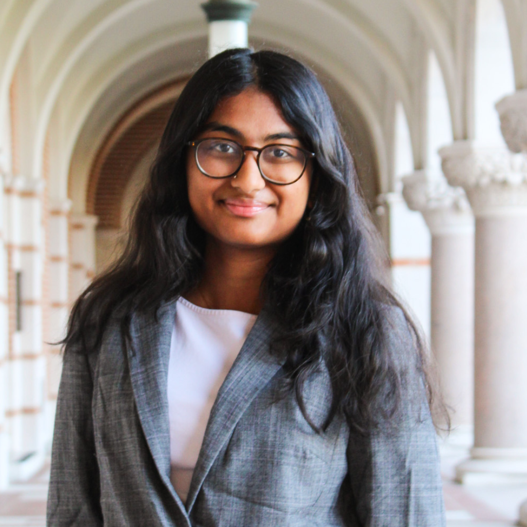 A female student is facing the camera and smiling. She has long, wavy black hair, glasses, and is wearing a gray blazer over a white blouse.