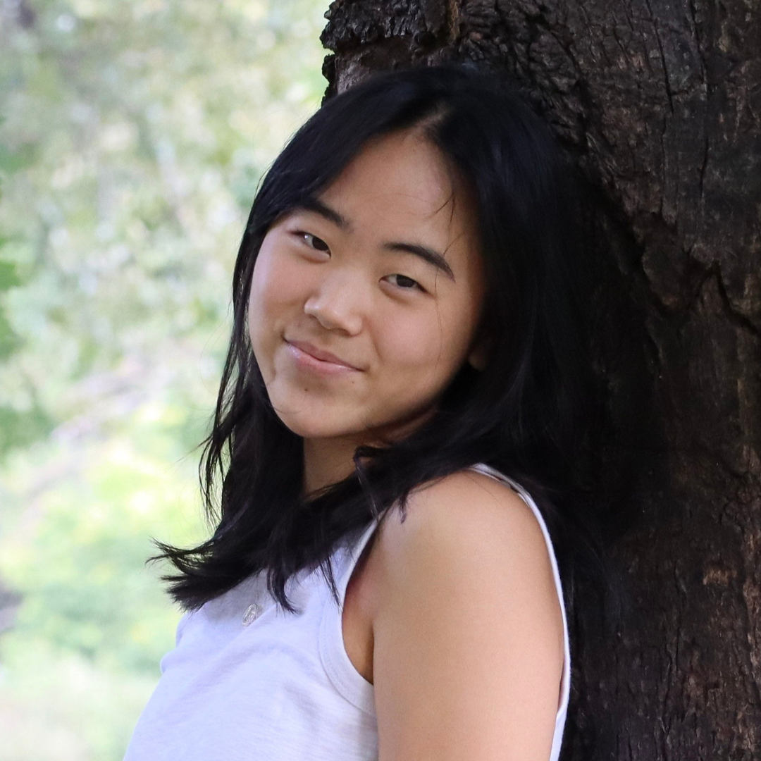 A female student with medium-length, straight black hair is leaning against a tree and wearing a pale tank top. She has a slight smile as she looks into the camera.