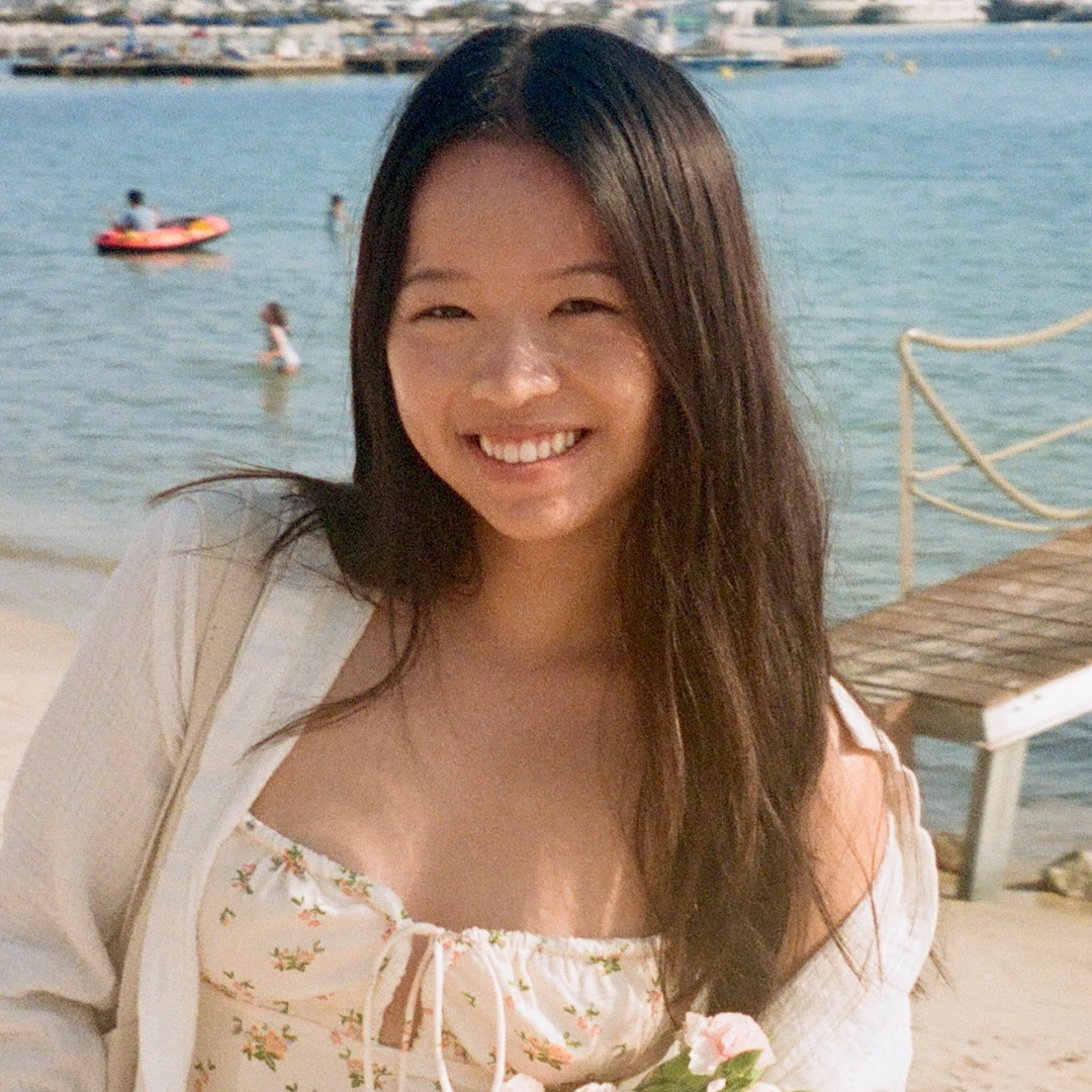 A female student with long, brown hair is smiling at the camera. She is wearing a white blouse with flowers on it and standing on a beach