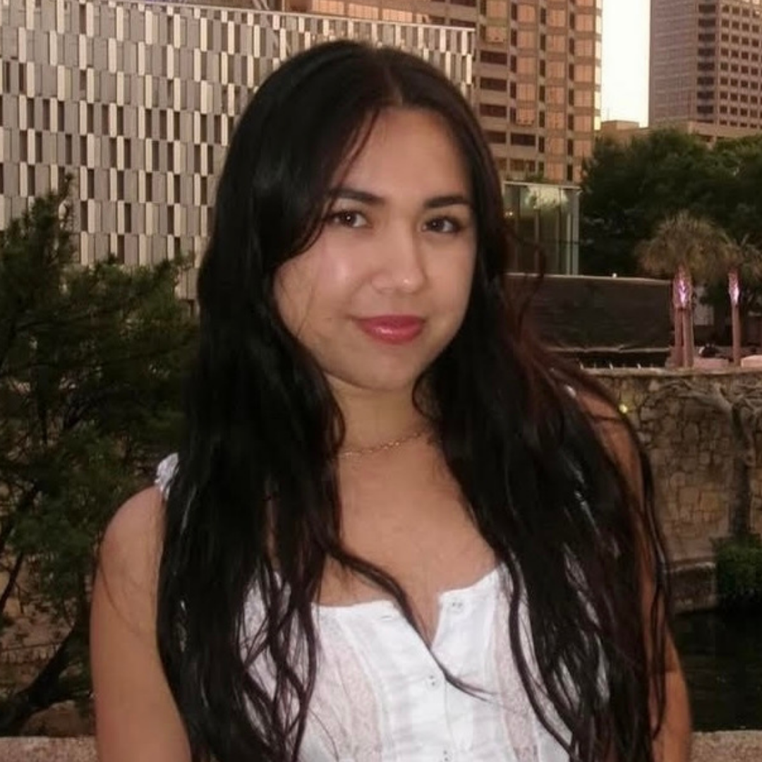 A female student with long, wavy black hair is smiling at the camera. She is wearing a white tank top and standing in front of a city skyline