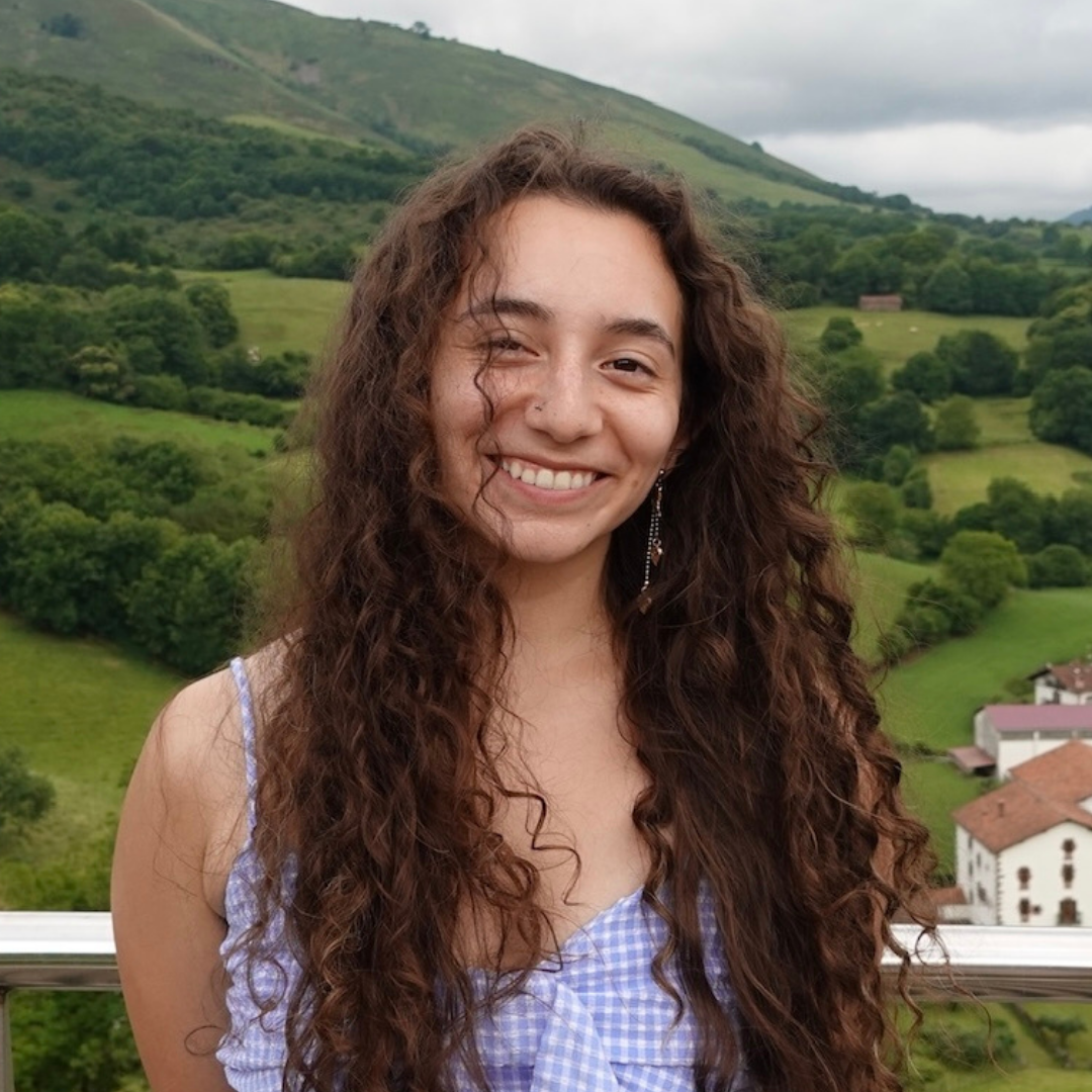 A female student with long, curly brown hair is smiling at the camera and wearing a gingham tanktop