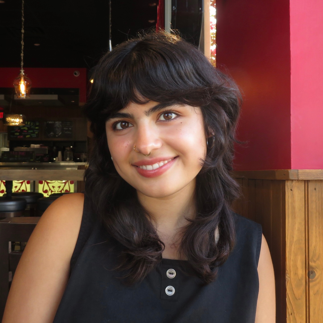 A female student with brown eyes is smiling at the camera. She has medium length, wavy, dark brown hair with bangs. She is also wearing a black tank top.
